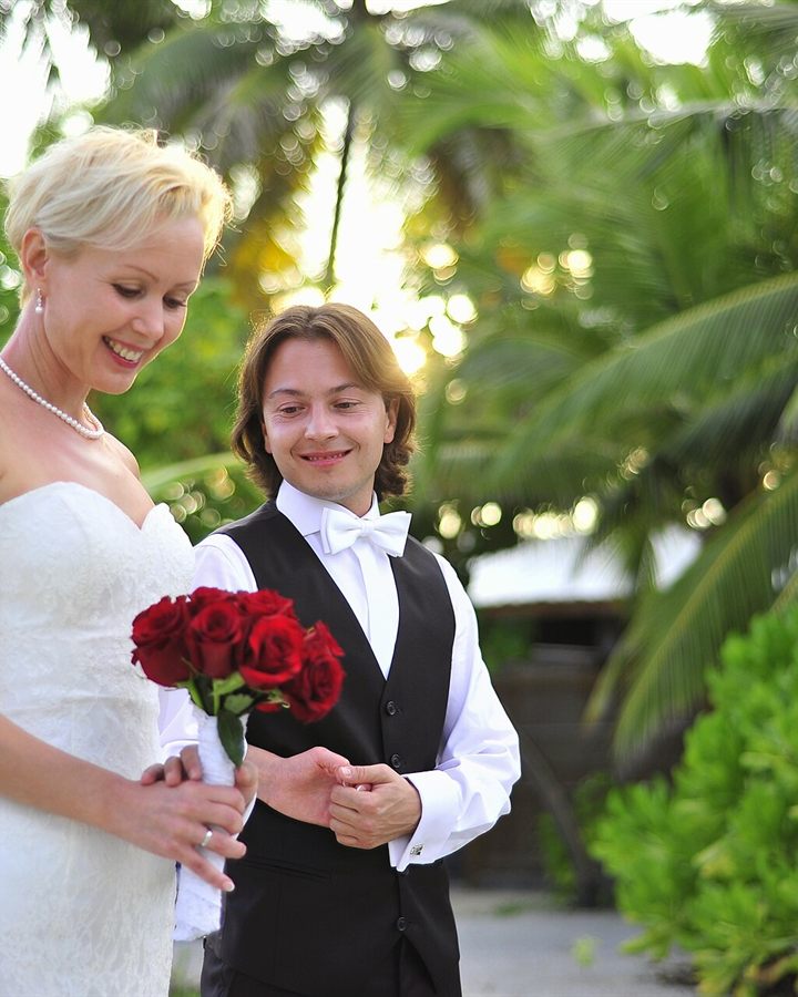 Bride and groom holding red roses beside palm trees at a tropical wedding